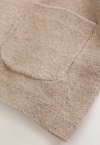 Close-up of a beige knitted fabric with a textured patch pocket and ribbed hem.
