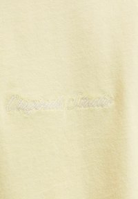 Απαλό κίτρινο ύφασμα με διακριτική κεντημένη καλλιγραφία που αναγράφει «Originals Studio» με νήμα στο ίδιο χρώμα, πάνω σε μια απαλή υφή.