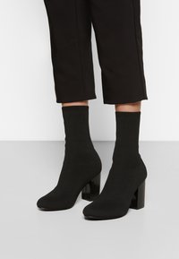 Botines de tobillo negros con una parte superior de tejido suave y elástico y tacones cuadrados. Presentan un cuello acanalado ajustado para un ajuste seguro.