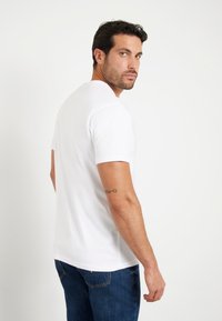 T-shirt blanc à manches courtes avec col rond, texture lisse et coupe ajustée, porté avec un jean en denim bleu. Tatouage remarquable sur le bras gauche.