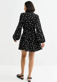 Schwarzes Polka-Dot-Kleid mit langen Ärmeln, tailliertem Schnitt und ausgestelltem Rock; aus leichtem Stoff gefertigt, mit weißen Polka-Punkten in unterschiedlichen Größen.