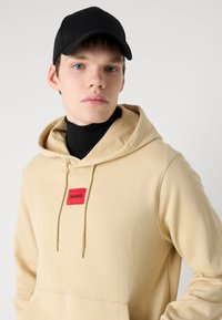 Sudadera beige con un parche rectangular rojo que dice "HUGO", cuello con cordón y una textura suave, combinada con una gorra negra.