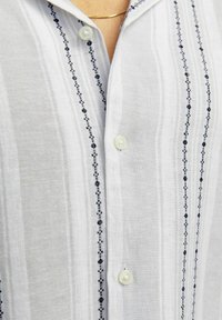 Chemise blanche à manches longues avec un motif rayé subtil et des accents brodés bleus au centre, fabriquée dans un tissu texturé et léger.