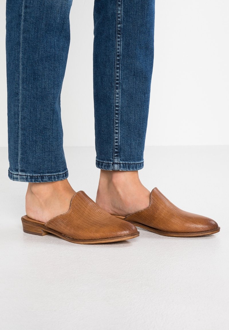 Personne portant un jean bleu et des mules en cuir tressé marron à talons en bois bas, posée sur un sol blanc.