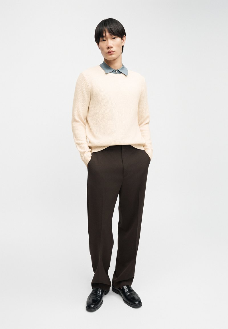 Man wearing beige sweater, gray collared shirt, dark brown trousers, and black leather shoes standing with hands in pockets.