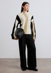 Pull en maille crème, noir et brun avec des motifs abstraits ; coupe oversize. Le mannequin tient un sac en cuir noir et porte un pantalon noir ample.