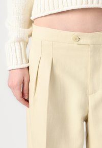 Pantalons plissés de couleur crème en tissu texturé, dotés d'une fermeture à un bouton et de poches latérales. Vue recadrée montrant la taille.