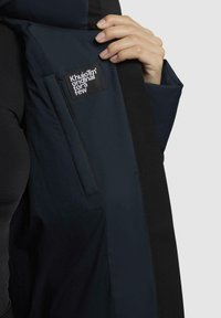 Giacca blu navy caratterizzata da una trama liscia, una tasca laterale e un'etichetta nera con testo bianco. Il design include un colletto e spalle a contrasto.