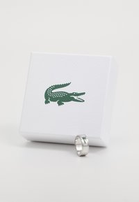 Sidabrinis žiedas su blizgiu paviršiumi, ant kurio pavaizduotas žalias krokodilo logotipas, pastatytas šalia tekstūruotos baltos dėžutės su tuo pačiu logotipu.