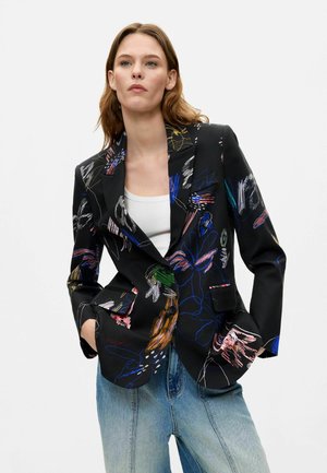 Donna che indossa un blazer nero con motivi floreali astratti colorati, top bianco e jeans azzurri chiari, in piedi su sfondo bianco.