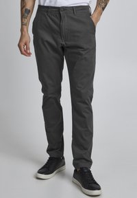 Mørkegrå chinos laget av mykt stoff, med rett ben-design, frontlommer og knappestang. Brukt med svarte joggesko.