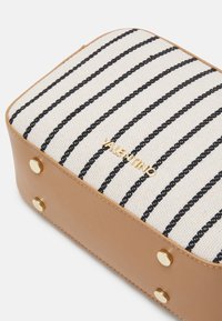 Obdélníková taška s plátěným vrškem, který má béžové a černé pruhy. Dno je z hnědé kůže s zlatými hroty a logem.