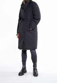Persoon in een lange zwarte gewatteerde jas met een riem, zwarte skinny jeans en zwarte chunky leren laarzen staand op een witte achtergrond.