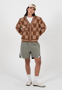 Femme portant un cardigan à carreaux marron, un short gris, des chaussettes blanches, des mocassins et une casquette beige, debout avec les mains dans le dos sur un fond blanc.