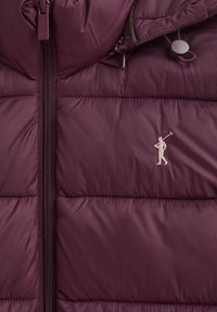 Zbliżenie na bordo pikowaną kurtkę z zamkiem błyskawicznym i haftowanym logo golfisty na piersi, wyposażoną w regulowany sznurek kaptura.