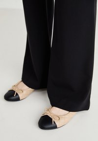 Balerini cu un exterior bej matlasat, vârf negru și detaliu cu fundiță; purtate cu pantaloni largi negri. Textură netedă și design simplu.