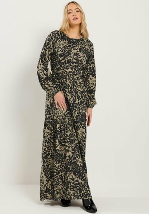 Blonde vrouw die een lange, loszittende jurk draagt met een zwart-olijfgroen patroon en lange mouwen, staand tegen een effen lichte achtergrond.