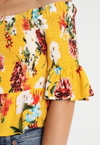 Top con hombros descubiertos en amarillo con estampado floral, que presenta textura fruncida y mangas con volantes. Los colores contrastantes incluyen rojo, blanco y azul.