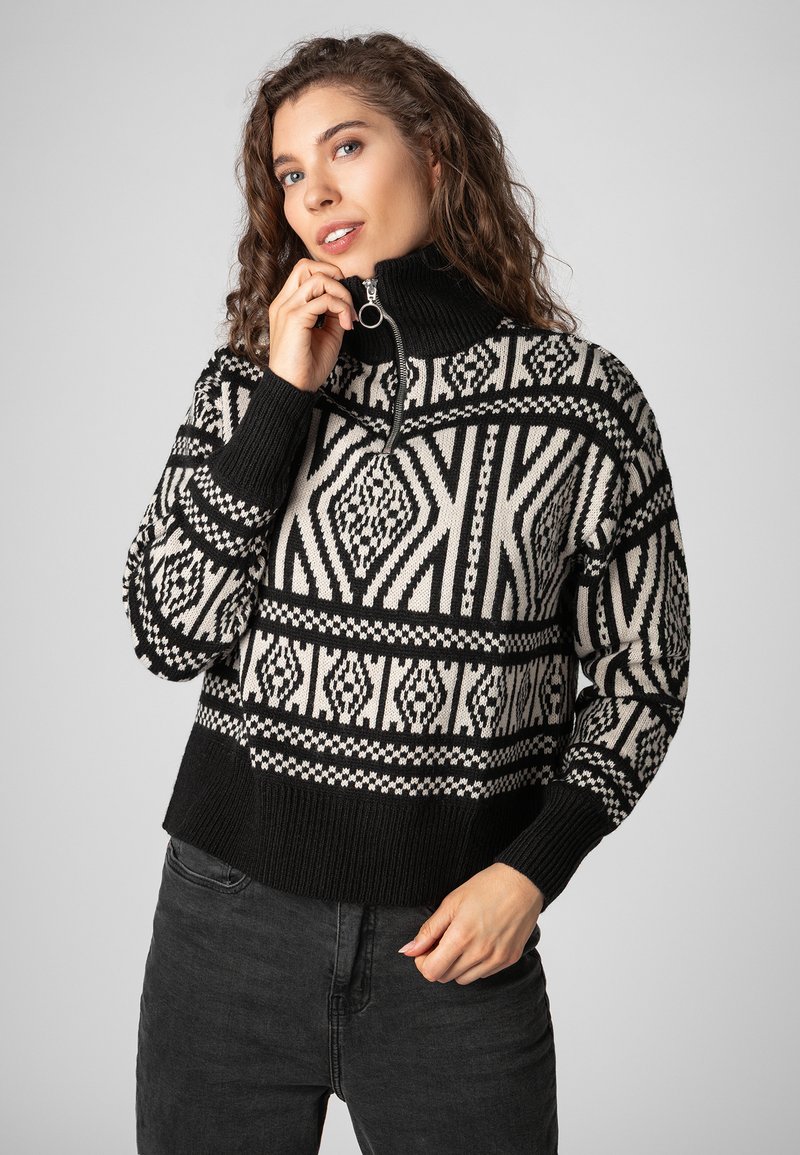 Schwarzer und cremefarbener Pullover mit Muster, hohem Kragen und halber Reißverschluss, der geometrische Designs in gestrickter Textur aufweist.