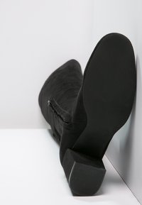 Stivale in camoscio nero fino a metà polpaccio con tacco a blocco, cerniera laterale e suola strutturata, posato su una superficie bianca contro un angolo di muro bianco.