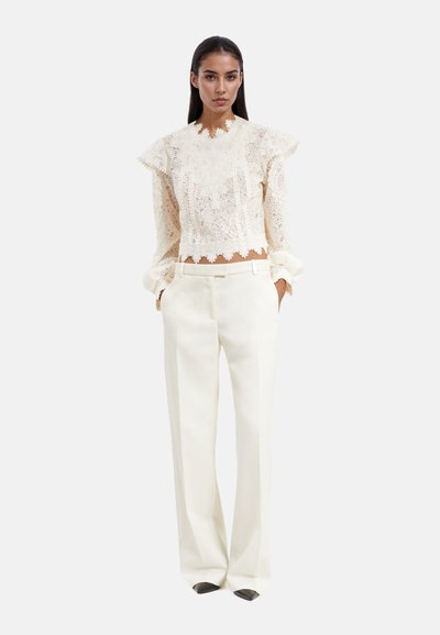 Blouse blanche en dentelle avec un col et des poignets volantés, associée à un pantalon crème taille haute et ajusté. Texture lisse, design raffiné.