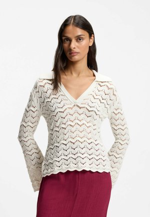 Vrouw met lang donker haar die een wit gebreide top met lange mouwen en zigzagpatroon draagt en een diepe V-hals, gecombineerd met een donkerroze rok.