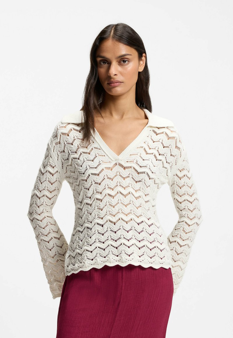 Femme aux longs cheveux foncés portant un haut blanc en maille à manches longues avec un motif chevron et un col en V profond, associé à une jupe rose foncé.