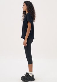 Zwarte katoenen T-shirt met korte mouwen, gecombineerd met zwarte leggings. Draag het met zwarte sneakers en witte sokken, gezien van opzij.