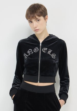 Giovane con capelli corti castani che indossa un hoodie nero in velluto corto con testo in strass e pantaloni abbinati, in piedi davanti a uno sfondo bianco.