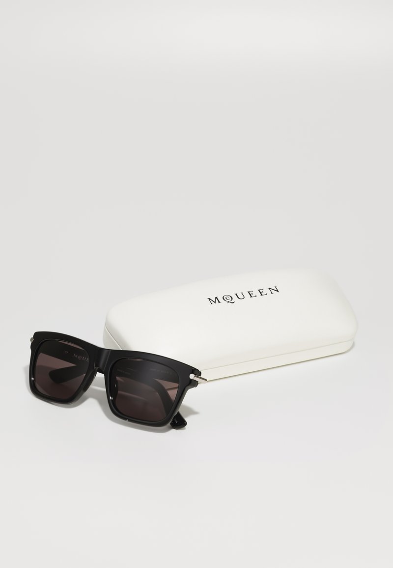 Occhiali da sole neri a forma quadrata con lenti scure e dettagli argentati, posizionati accanto a un astuccio rigido bianco con il logo "MQUEEN" stampato in nero.
