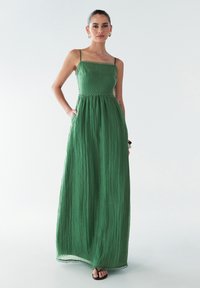 Grünes Maxikleid mit dünnen Trägern, strukturiertem Stoff, elastischem Bund und Spitzenbesatz am Saum. Modell posiert mit den Händen in den Taschen.