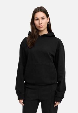 Schwarzer Hoodie aus weichem Stoff, mit einer frontalen Tasche und einer verstellbaren Kapuze mit Kordelzug. Kombiniert mit passenden schwarzen Hosen.