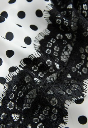 Czarna koronkowa lamówka na białej tkaninie z czarnymi kropkami. Tkanina ma gładką fakturę; koronka zawiera kwiatowe wzory i postrzępione krawędzie.