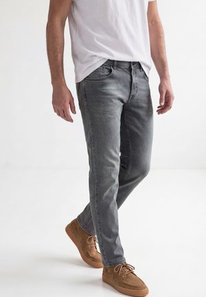 Mand iført falmede grå jeans, hvid t-shirt og brune afslappede sko, stående med det ene ben krydset over det andet på en hvid baggrund.