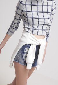 Top corto de manga larga de cuadros gris combinado con shorts de mezclilla desgastados. Un suéter blanco atado a la cintura añade un detalle en capas.