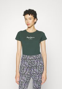Žalia medvilninė marškinė su trumpomis rankovėmis, su baltu "Pepe Jeans" logotipu. Derinama su juodomis kelnėmis su gėlių raštu purpuriniais ir baltais akcentais.