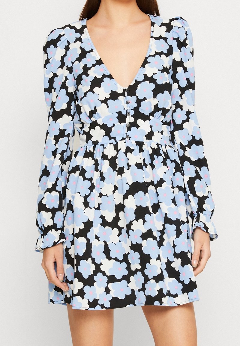 Femme portant une robe noire avec un imprimé floral bleu et blanc, manches longues, boutons sur le devant et un profond décolleté en V.