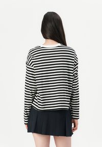 Donna con lunghi capelli scuri che indossa una maglietta a righe bianco e nera con maniche lunghe e una gonna plissettata nera, voltata di spalle su uno sfondo bianco.