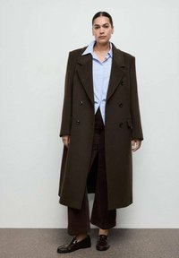 Brązowy wełniany płaszcz z podwójnym rzędem guzików, w połączeniu z jasnoniebieską, w paski koszulą i ciemnobrązowymi spodniami o szerokich nogawkach. Nosi czarne mokasyny.