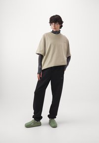 Přehnaně velké béžové bavlněné tričko, černé volné kalhoty a zelené slip-on boty s výřezy, umístěné na jednolitém bílém pozadí.