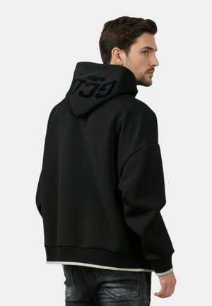 Uomo che indossa una felpa con cappuccio nera con la scritta "GCDS" in grandi lettere sul retro del cappuccio, abbinata a jeans scuri, visto di lato.