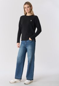 Maglione nero lavorato a maglia con scollo rotondo, abbinato a jeans ampi azzurri e sneakers bianche. Design semplice, vestibilità casual.