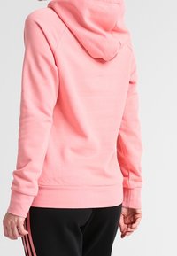 Rosa hoodie med avslappnad passform, med dragsko i huvan, långa ärmar och ribbade ärmslut. Kombineras med svarta byxor med rosa ränder.