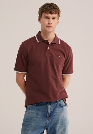 Giovane con capelli ricci che indossa una polo bordeaux con rifiniture bianche e jeans blu, in piedi davanti a uno sfondo chiaro e uniforme.