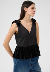 Haut sans manches noir avec un décolleté en V, featuring un ourlet en velours à volants et des accents de nœud en tissu sur les épaules, associé à un jean en denim bleu.