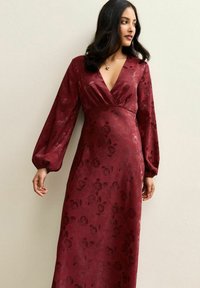 Bordowa sukienka kopertowa w kwiatowy wzór, z długimi bufkowatymi rękawami i głębokim dekoltem w kształcie litery V. Wykonana z gładkiej tkaniny, dopasowana w talii, swobodnie opadająca aż do podłogi.