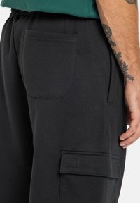 Schwarze Cargo-Hosen aus weichem Material, mit einer Gesäßtasche und einer flachen Vorderseite, die auffällige seitliche Cargo-Taschen aufweist. Flache Nähte und elastischer Bund.