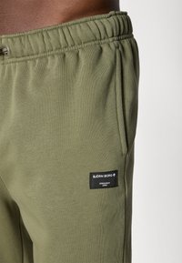 Olivza zaņģu bikses ar elastīgu jostu, sānu kabatām un izšūtu melnu birku, kurā redzams "BJÖRN BORG" un "STOCKHOLM 1956."