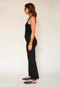 Top noir sans manches, pantalons noirs ajustés et chaussures noires avec des accents argentés. Bracelets en or au poignet. Vue de profil contre un fond neutre.