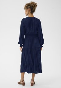 Tumši zils garām piedurknēm kleita ar vairāku līmeņu ruffles dizainu. Smocked viduklis un teksturēts audums. Valkāta ar blokveida papēžiem sandalēm.